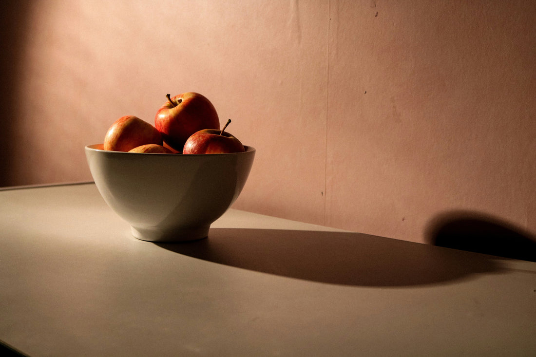 A dark, beige background with a fruit bowl sitting in front containing red apples. 