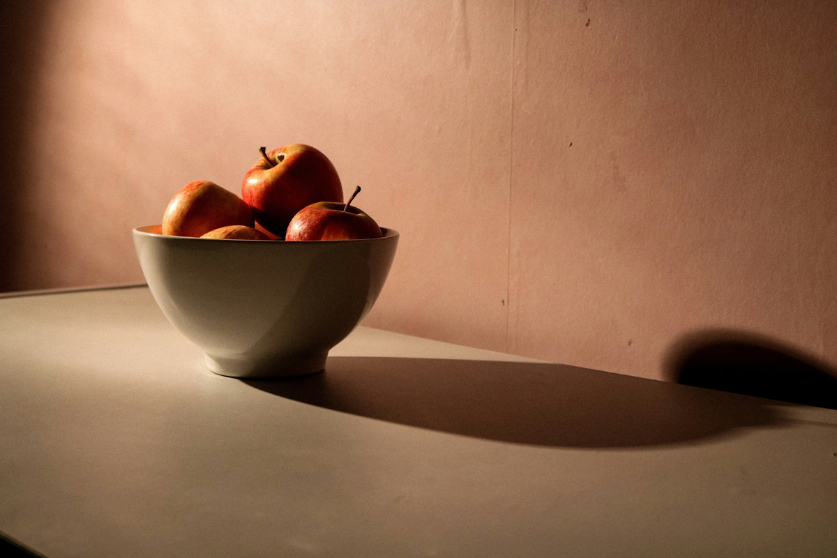 A dark, beige background with a fruit bowl sitting in front containing red apples. 